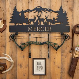 May include: Black metal wall art featuring a mountain range, trees, and deer silhouettes, with the name "MERCER" below. A green and black compound bow hangs below, with a framed wolf drawing and a quiver of arrows.