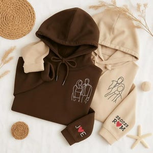 Könnte beinhalten: Zwei Kapuzenpullover, einer braun und einer beige, mit gestickten Strichzeichnungen von Paaren. Der braune Pullover hat "J♡E" am Bündchen. Der beige Pullover hat "23.07.23 R♡M" am Bündchen. Beide haben Kordelzüge.