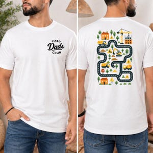 Può includere: T-shirt bianca con la scritta "Tired Dads Club" in nero sul davanti. Il retro della maglietta presenta una grafica colorata di una strada con veicoli da costruzione, case, alberi e una gru.