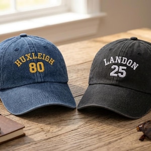 Peut inclure: Deux casquettes de baseball, une bleu denim et une gris anthracite, sont posées sur une surface en bois. La casquette bleue porte la mention "HUXLEIGH 80" brodée en or. La casquette grise affiche "LANDON 25" en blanc. Un carnet, un stylo et des lunettes de soleil sont également visibles.