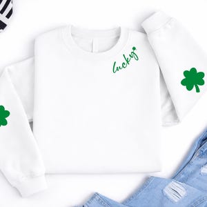Puede incluir: Sudadera blanca de manga larga con un trébol verde en la manga izquierda y un trébol verde y la palabra "Lucky" en la parte delantera. La sudadera se combina con unos vaqueros azules. Se ve un sombrero a rayas blancas y negras.