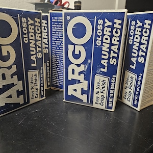 Puede incluir: Cuatro cajas azules y blancas de almidón para ropa Argo Gloss están dispuestas sobre una superficie oscura. Cada caja tiene el logotipo de Argo, información del producto y el texto "Easy To Use... Crisp Finish". Cada caja contiene 454 g.