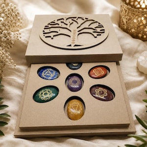 May include: A light brown wooden box with a laser-cut tree of life design on the lid, containing seven oval-shaped polished stones. Each stone features a unique gold-colored chakra symbol engraved on its surface, arranged in a specific pattern.