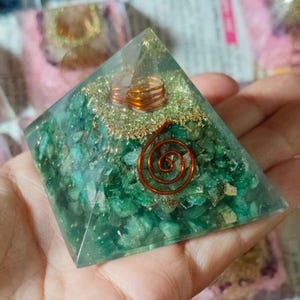 May include: A clear resin pyramid with green stone chips and copper wire accents. The pyramid features a copper spiral and gold-colored flakes. The pyramid is held in a hand.
