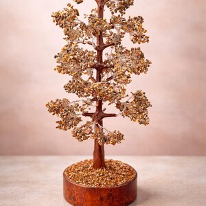 May include: A decorative tree sculpture with a brown trunk and branches, adorned with small, light brown stones. The tree sits in a round, wooden base filled with similar stones. The background is a soft, muted pink.
