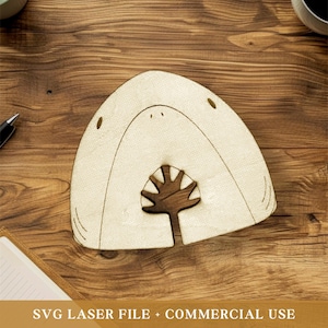 Puede incluir: Un posavasos de madera con forma de tiburón, con un exterior beige claro y un interior marrón oscuro. El posavasos presenta un diseño recortado de la boca y los dientes de un tiburón. El texto "SVG LASER FILE + COMMERCIAL USE" está en la parte inferior.