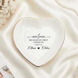 Puede incluir: Un plato de cerámica blanco en forma de corazón con borde dorado. El plato tiene el texto "we love because he first loved us 1 John 4:19" y los nombres "Ethan" y "Chloe". Un regalo de boda.