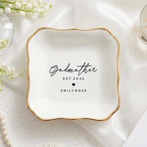 May include: A white ceramic trinket dish with a gold rim, featuring the inscription "Godmother EST 2026 EMILY ROSE" in black script. The dish is square with wavy edges, and sits on a white satin surface.