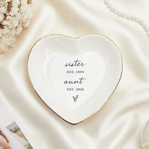 Peut inclure: Un vide-poche en céramique blanc en forme de cœur avec un bord doré, portant les mots "sister" et "aunt" avec des dates. Le plat est posé sur une surface en satin blanc, avec une photo de mariage et des fleurs à proximité.