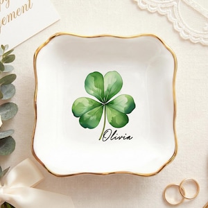May include: White ceramic trinket dish with a gold rim, featuring a watercolor shamrock design and the name "Olivia". The dish is square with wavy edges. A "Happy Engagement" card and flowers are in the background.