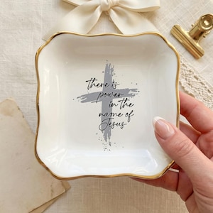 Puede incluir: Un plato de cerámica blanco con borde dorado y el texto "there is power in the name of Jesus" en escritura negra. Un diseño de cruz gris está detrás del texto. El plato está sostenido por una mano.