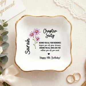 Könnte beinhalten: Eine weiße Keramik-Schmuckschale mit goldenem Rand, mit dem Text "Chapter Sixty" und einem floralen Design. Die Schale enthält den Namen "Sarah" und eine Geburtstagsbotschaft. Ein Blumenstrauß und eine Karte mit der Aufschrift "Happy Engagement" sind im Hintergrund.
