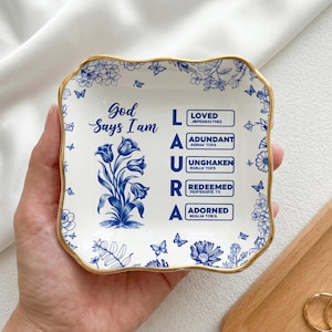 Puede incluir: Un plato de cerámica blanco con un diseño floral azul y ribete dorado. El plato presenta el texto "Dios dice que soy" y el nombre "LAURA" con palabras asociadas. Mariposas y flores decorativas adornan los bordes.