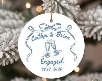 May include: White round ceramic ornament with a blue design. The ornament features the names "Caitlyn & Brian", two champagne flutes, and the word "Engaged" with the date 08.17.2026. A gold string is attached for hanging.