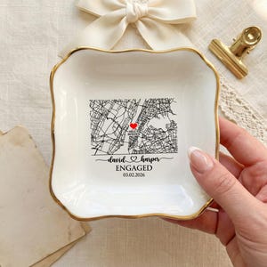 Puede incluir: Plato de cerámica blanca para baratijas con borde dorado. El plato presenta un diseño de mapa en blanco y negro con un corazón rojo, los nombres "David & Harper" y la palabra "ENGAGED" con la fecha 03.02.2026.