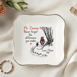 May include: A white, square trinket dish with a gold rim. The dish features a black and red illustration of two bears walking on a path, with text that reads "Mr. Davies, Never forget the difference you make."