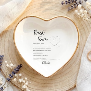 May include: A white heart-shaped ceramic trinket dish with a gold-painted rim. The centre features black text reading Best Team, a definition of the term, and the name Olivia, accompanied by a small dashed-line heart graphic.