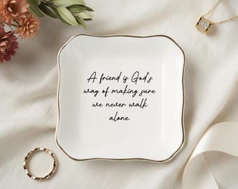 May include: A white, square trinket dish with a gold rim and the quote, "A friend is God's way of making sure we never walk alone." A gold ring and necklace are also visible.