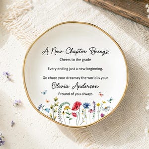 May include: A round white ceramic trinket dish with a gold-painted rim. The center features black text reading A New Chapter Beings, Cheers to the grade, Every ending just a new beginning, Go chase your dreamay the world is your, Olivia Anderson, Proud of you always, and a colorful floral and butterfly illustration.