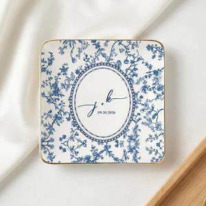 Op de afbeelding: Vierkante keramische sieradenschaal met een blauw bloemenpatroon en gouden rand. Het midden heeft een ovaal ontwerp met de initialen "J + K" en de datum "09.20.2026" in een scriptlettertype. De schaal ligt op een wit oppervlak.