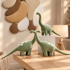 Pode incluir: Três dinossauros de pelúcia verde-sálvia com pescoços e caudas longos. Cada dinossauro apresenta uma fileira de pequenos pontos redondos de feltro branco nas costas. Os brinquedos estão sobre uma superfície de madeira, um sobre um bloco.
