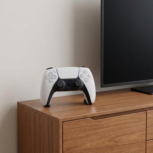 May include: A white and black PlayStation 5 DualSense wireless controller. The controller is on a wooden cabinet. The controller has a white face with black accents and buttons. The controller has a symmetrical design with two analog sticks and a directional pad.