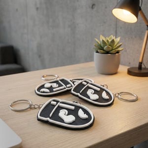 May include: Several black and white keyrings with a distinctive design, featuring a black base and white raised details. The keyrings are displayed on a wooden surface, with a small plant and a desk lamp in the background.