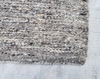 Puede incluir: Primer plano de una alfombra texturizada de tonos neutros. La alfombra presenta una mezcla de fibras grises, blancas y marrones, creando una apariencia moteada. Los bordes están deshilachados, lo que añade encanto rústico a la alfombra. La alfombra es probablemente de lana o material similar.