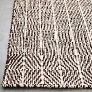 Può includere: Un tappeto rettangolare intrecciato caratterizzato da un motivo geometrico ripetuto di rombi color marrone scuro e crema. Il tappeto presenta una superficie testurizzata con fibre di filato visibili e un bordo rifinito con una griglia.