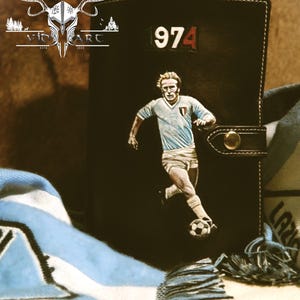Può includere: Diario in pelle nera con un'illustrazione di un calciatore e il numero "974". Il diario ha una chiusura a bottone ed è impreziosito da una sciarpa blu e bianca. Il logo "Vio Art" è visibile in alto a sinistra.