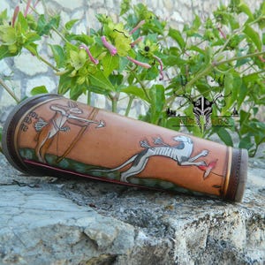 Könnte beinhalten: Ein zylindrischer, brauner Lederarmschutz mit handgemalten Illustrationen eines Jägers mit Pfeil und Bogen und eines laufenden Tieres. Der Armschutz hat eine dunkelbraune Umrandung und liegt auf einer Steinoberfläche.