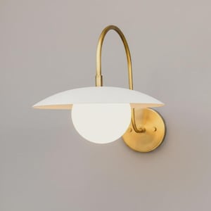 Puede incluir: Aplique de pared moderno con una pantalla blanca en forma de platillo y una bombilla esférica de vidrio esmerilado. La lámpara presenta un brazo curvo de color latón y una base circular.
