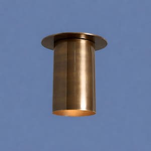 May include: A cylindrical, brass-colored light fixture with a circular top and a slightly flared base. The fixture has vertical grooves along its sides and emits a warm, inviting glow from within. The background is a solid blue.