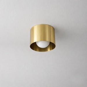 Könnte beinhalten: Eine goldfarbene, zylindrische Deckenleuchte. Die Leuchte hat einen abgerundeten, offenen Boden, der eine weiße, kugelförmige Glühbirne freilegt. Das Licht ist an einer weißen Decke montiert.