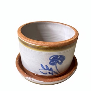 May include: A ceramic plant pot with a matching saucer. The pot is off-white with a brown rim and a blue floral design. The saucer is also brown. This handmade pottery is suitable for small plants.