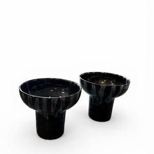May include: Two black ceramic bowls with a pedestal base. The bowls have a glossy finish with subtle streaks of white and gray. The design is modern and minimalist, suitable for serving or display.