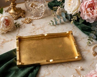 May include: A rectangular gold-toned tray with raised edges and a smooth, metallic finish. The tray features a flat base and a slightly ornate, structured border design, presenting a clean and reflective surface for display purposes.