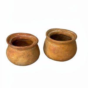 May include: Two small, round, terracotta-coloured clay pots with a textured, uneven surface and a wide, open mouth. Each vessel features a thick, rounded rim and a bulbous body, showcasing natural variations in the earthy brown and orange tones.