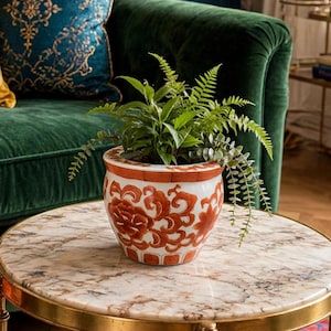 Peut inclure: Un pot de fleurs en céramique ronde présentant une base blanche avec un motif floral orange complexe et une bordure géométrique sur le rebord. Le pot contient une fougère verte luxuriante aux frondes délicates dépassant des côtés.