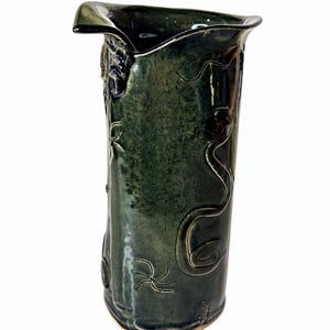 May include: A tall, dark green ceramic vase with a unique, asymmetrical rim. The vase features embossed, swirling designs and a textured surface. The handmade pottery piece is suitable for home decor.