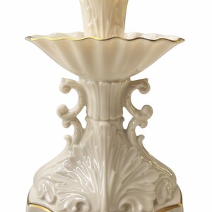 May include: A cream-colored porcelain candlestick holder featuring a central candle socket, a wide scalloped bobeche, and a decorative base with ornate leaf-like relief carvings. The edges of the bobeche and the base are trimmed with gold.
