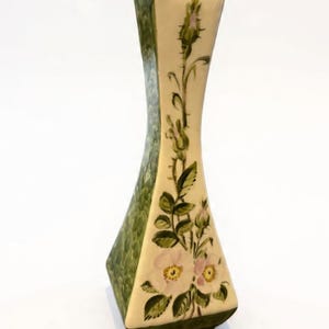 May include: A decorative ceramic vase with an hourglass shape. The vase has a cream-coloured central panel with pink and yellow floral designs and green leaves. The sides are textured green, and the base is dark green.