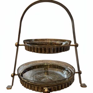 May include: Two-tiered serving stand with clear glass dishes and a decorative metal frame. The frame has a handle and is made of a gold-toned metal. The dishes have a scalloped edge.