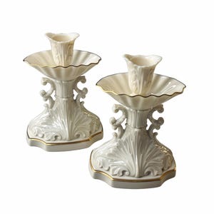 May include: A pair of off-white porcelain candle holders featuring a scalloped bobeche, a central fluted stem, and a wide base with embossed leaf motifs. Each piece is finished with a thin gold trim along the edges of the base and the bobeche.