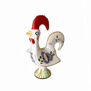 Puede incluir: Una figura de gallo de cerámica blanca con una cresta roja brillante, un pico amarillo y detalles florales pintados a mano en azul y verde. El gallo se apoya sobre una base circular con marcas verdes similares a hierba y tiene una cola curva.