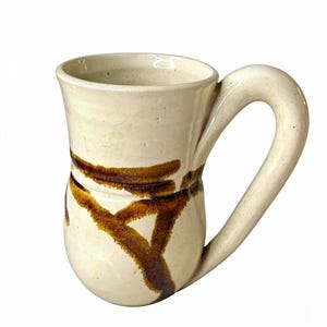 May include: A cream-colored ceramic mug with a speckled finish and a large, thick handle. The mug features a hand-painted, dark brown abstract brushstroke design that encircles the middle section, creating a distinct contrast against the light base.