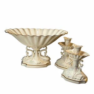 May include: A cream-colored ceramic centerpiece bowl with a scalloped rim and two matching candle holders. Each piece features ornate, molded scrollwork on the base and is finished with a thin gold trim along the edges of the bowl and stands.