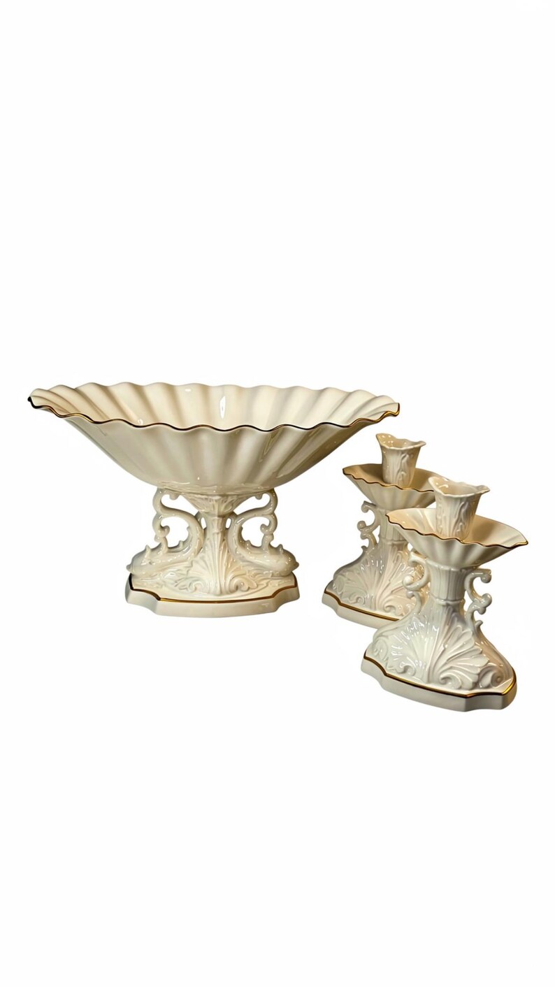 May include: A cream-colored ceramic centerpiece bowl with a scalloped rim and two matching candle holders. Each piece features ornate, molded scrollwork on the base and is finished with a thin gold trim along the edges of the bowl and stands.