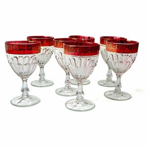 May include: A collection of seven clear wine glasses, each with a red rim and gold filigree detailing. The glasses feature a decorative, cut-glass pattern on the bowl, with clear stems and bases. Ideal for serving wine.