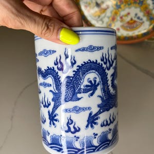 May include: A white and blue ceramic cup with a dragon design. The dragon is blue and wraps around the cup, with flames and clouds. The cup has a blue rim and base. A hand with yellow nail polish is holding the cup.